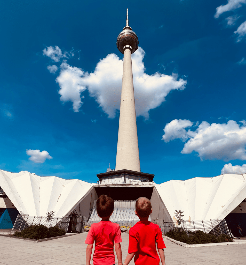 Zu sehen ist der Berliner Fernsehturm von der Seite des Neptunbrunnen. Es ist strahlend blauer Himmel mit ein paar Schäfchenwolken und zentral im Bild sind zwei Kinder, vermutlich beides Jungs, die beide ein knallrotes Shirt tragen und mit dem Rücken zum Betrachter auf den Berliner Fernsehturm blicken und sich bei der Hand nehmen.